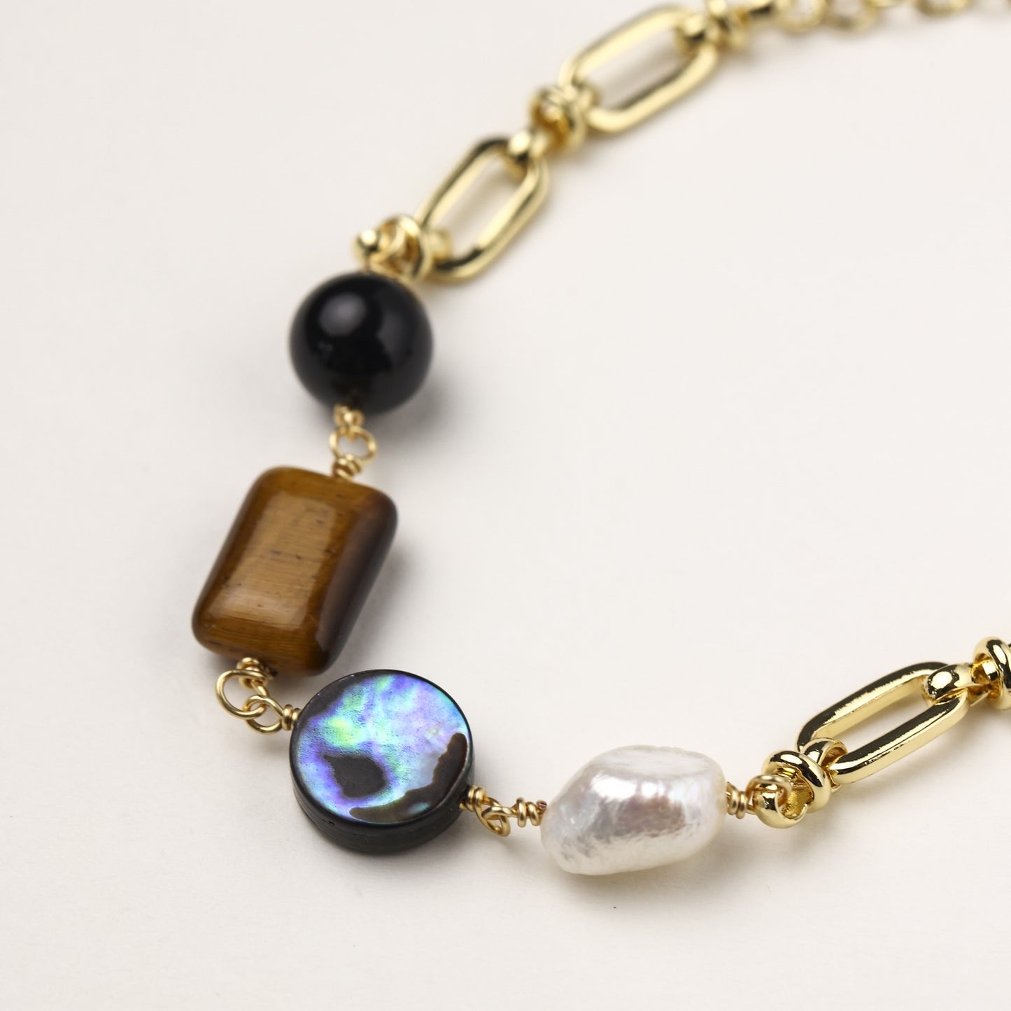 Gold-plated chain bracelet featuring tiger's eye, obsidian, freshwater pearls and abalone shell on white background