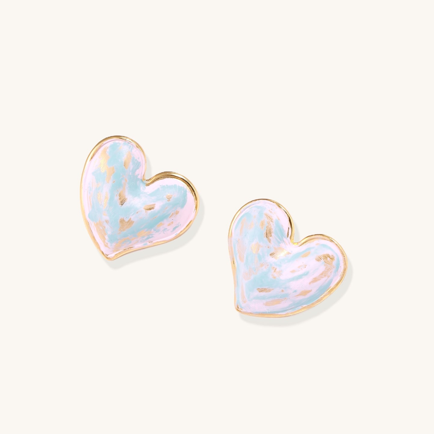Heart-shaped stud earrings with iridescent blue-pink enamel finish and gold-plated brass frames against cream background