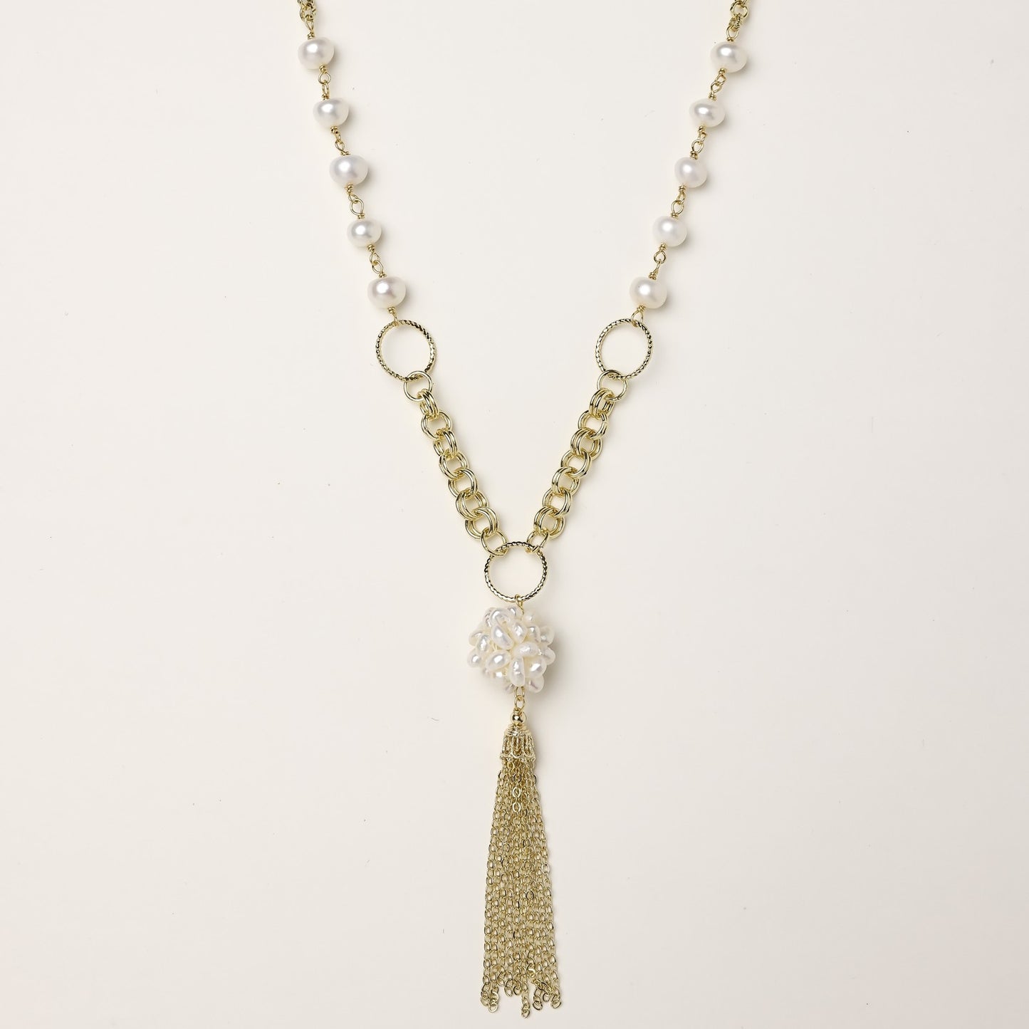 Layered gold-plated necklace with freshwater pearls, central pearl cluster, and chain tassel on cream background.