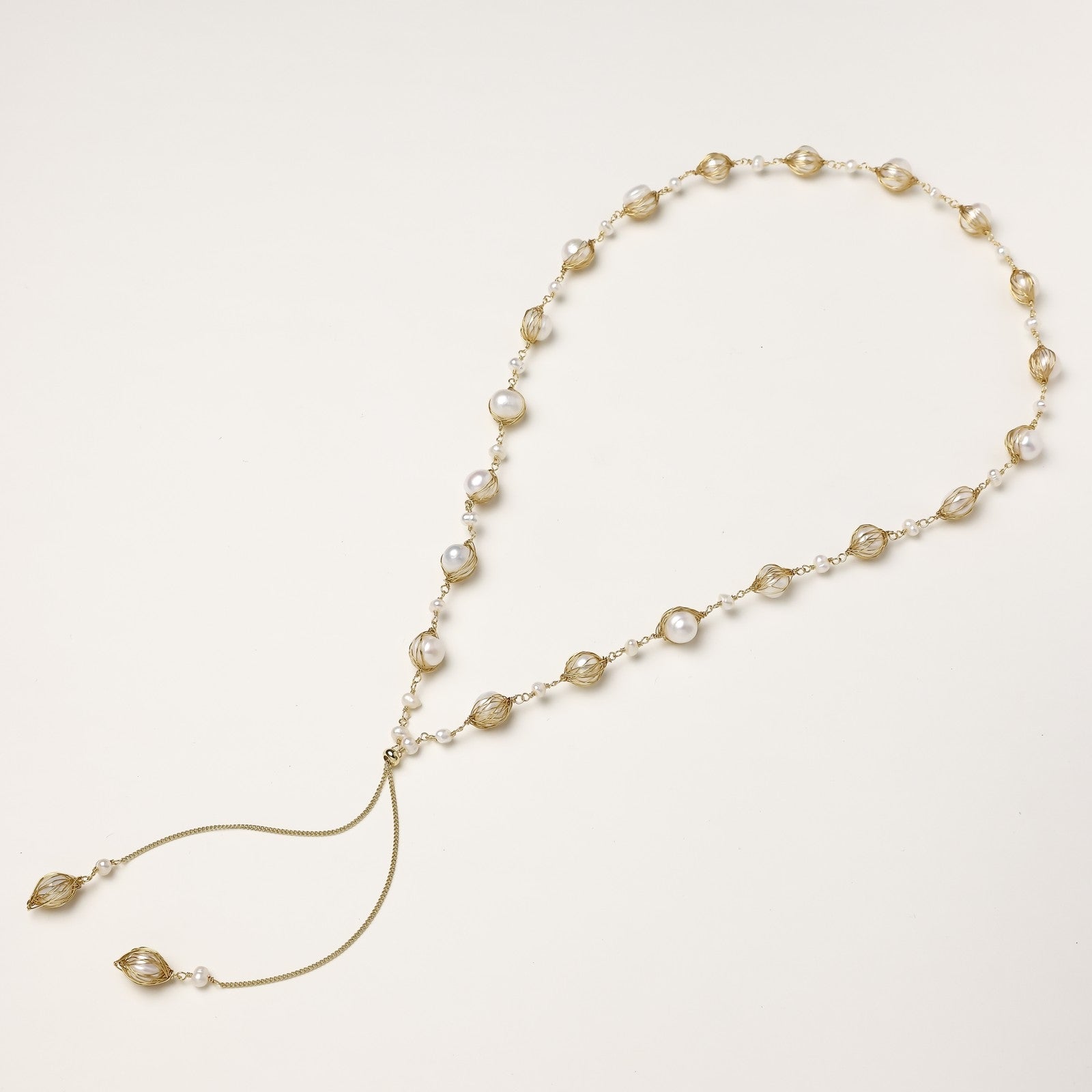 Long gold-plated lariat necklace with wire-wrapped freshwater pearls and chain tassel detail on cream background.