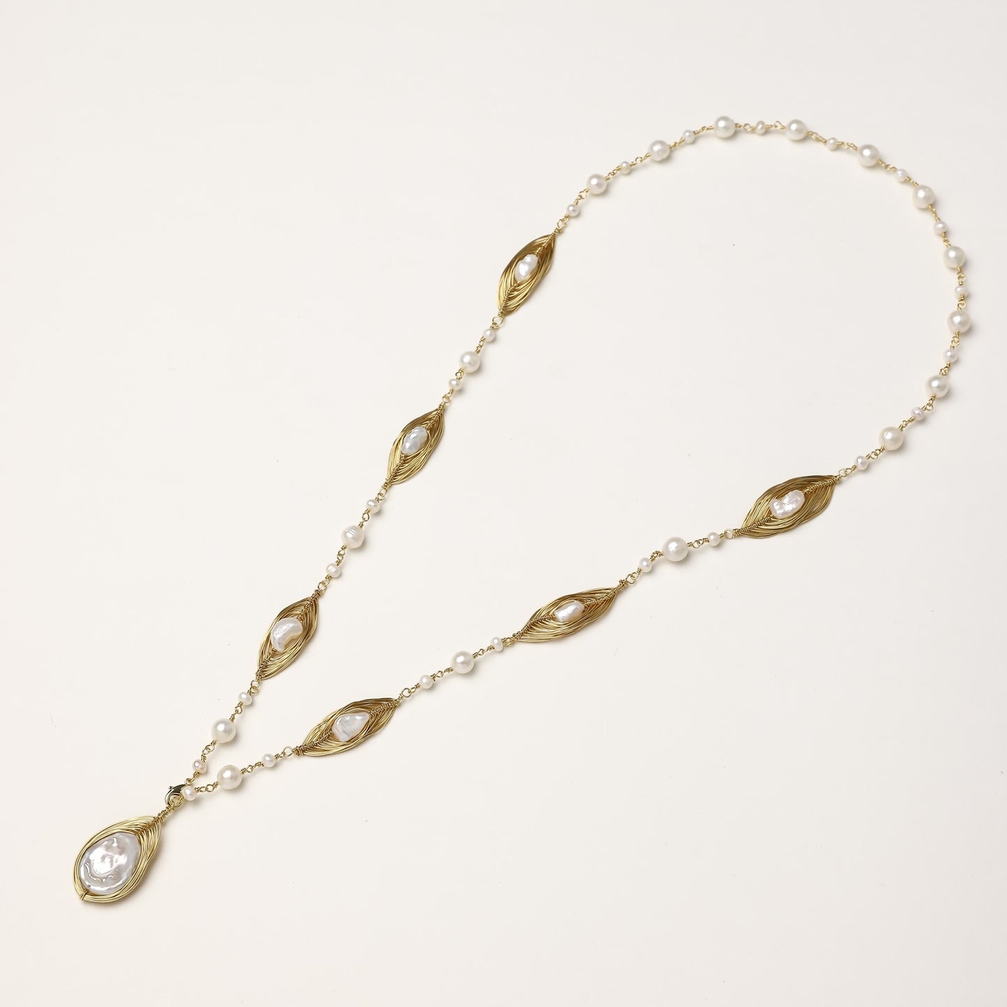 Long gold-plated necklace with eye-shaped elements, freshwater pearls, and baroque pearl pendant on neutral background.