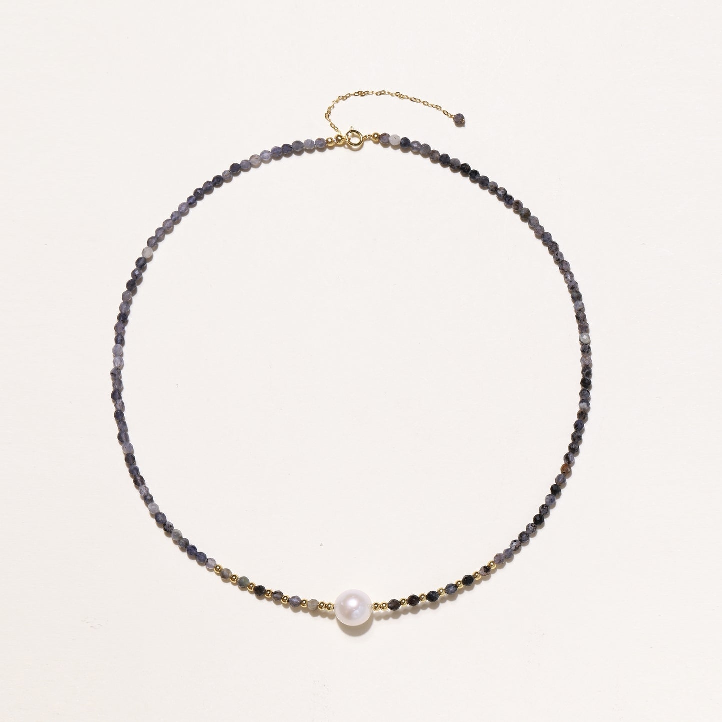 Delicate beaded necklace with violet iolite stones, gold spacers, and central white freshwater pearl on neutral background.