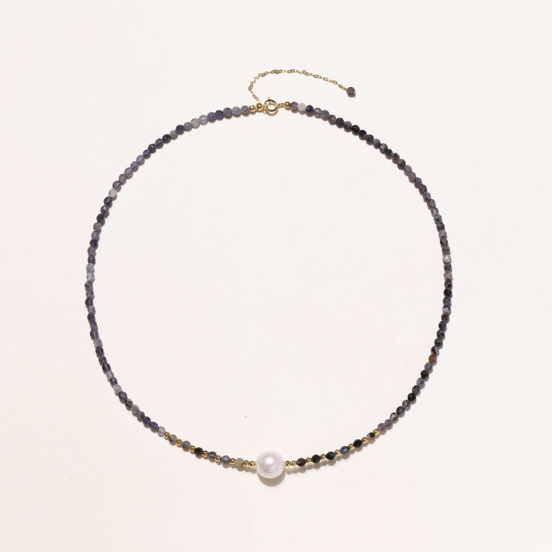Delicate beaded necklace with violet iolite stones, gold spacers, and central white freshwater pearl on neutral background.