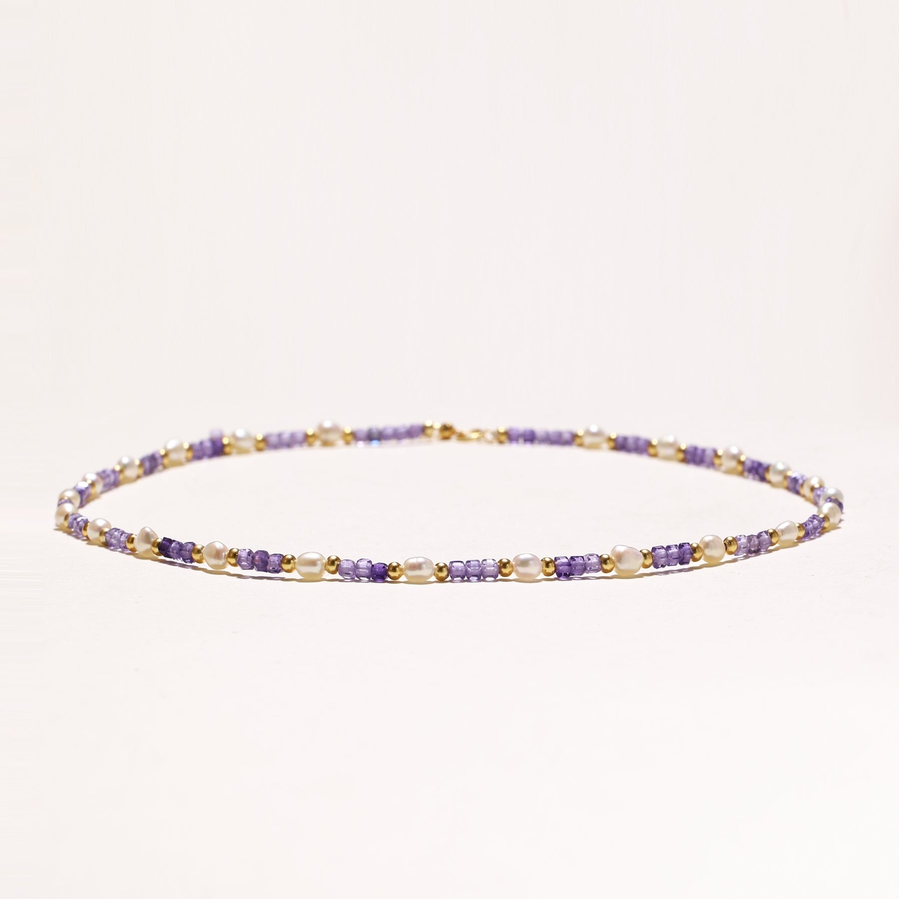 Delicate beaded necklace with alternating purple zirconia stones, white freshwater pearls, and gold-plated spacers on cream background.