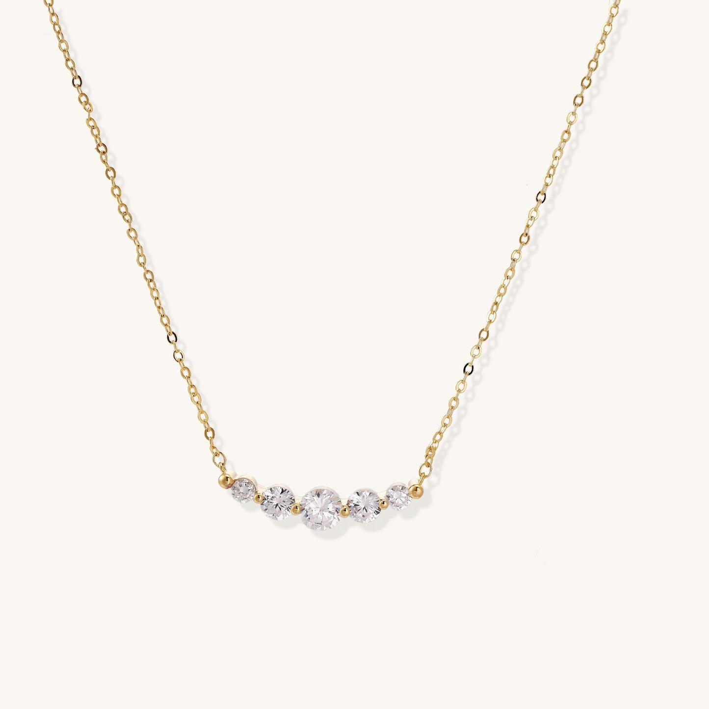 Delicate 14K gold plated necklace with curved arc of sparkling zirconia stones on cream background.