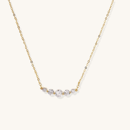 Delicate 14K gold plated necklace with curved arc of sparkling zirconia stones on cream background.