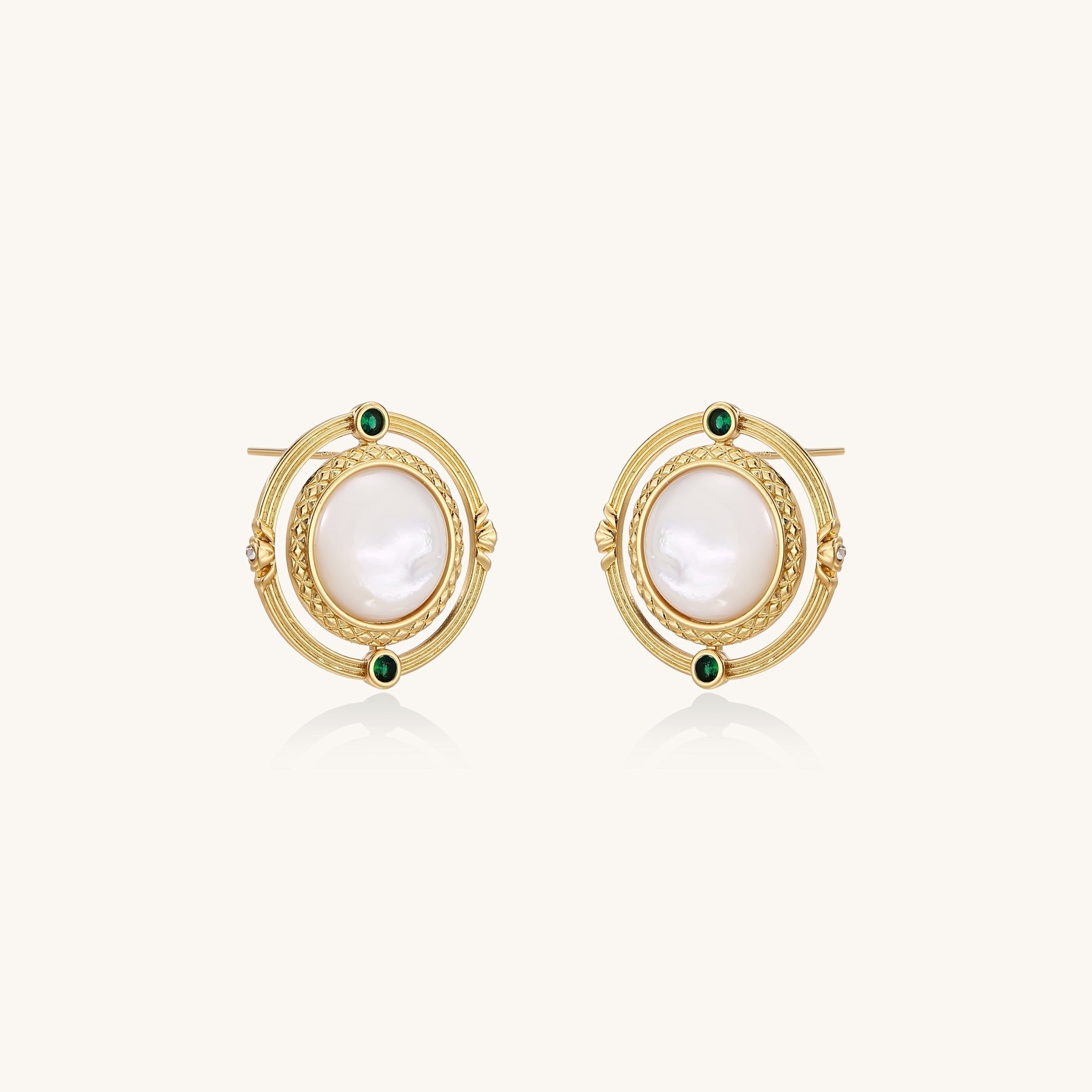 Close-up of gold-plated circular earrings with white mother-of-pearl centers and green zirconia accents on cream background.