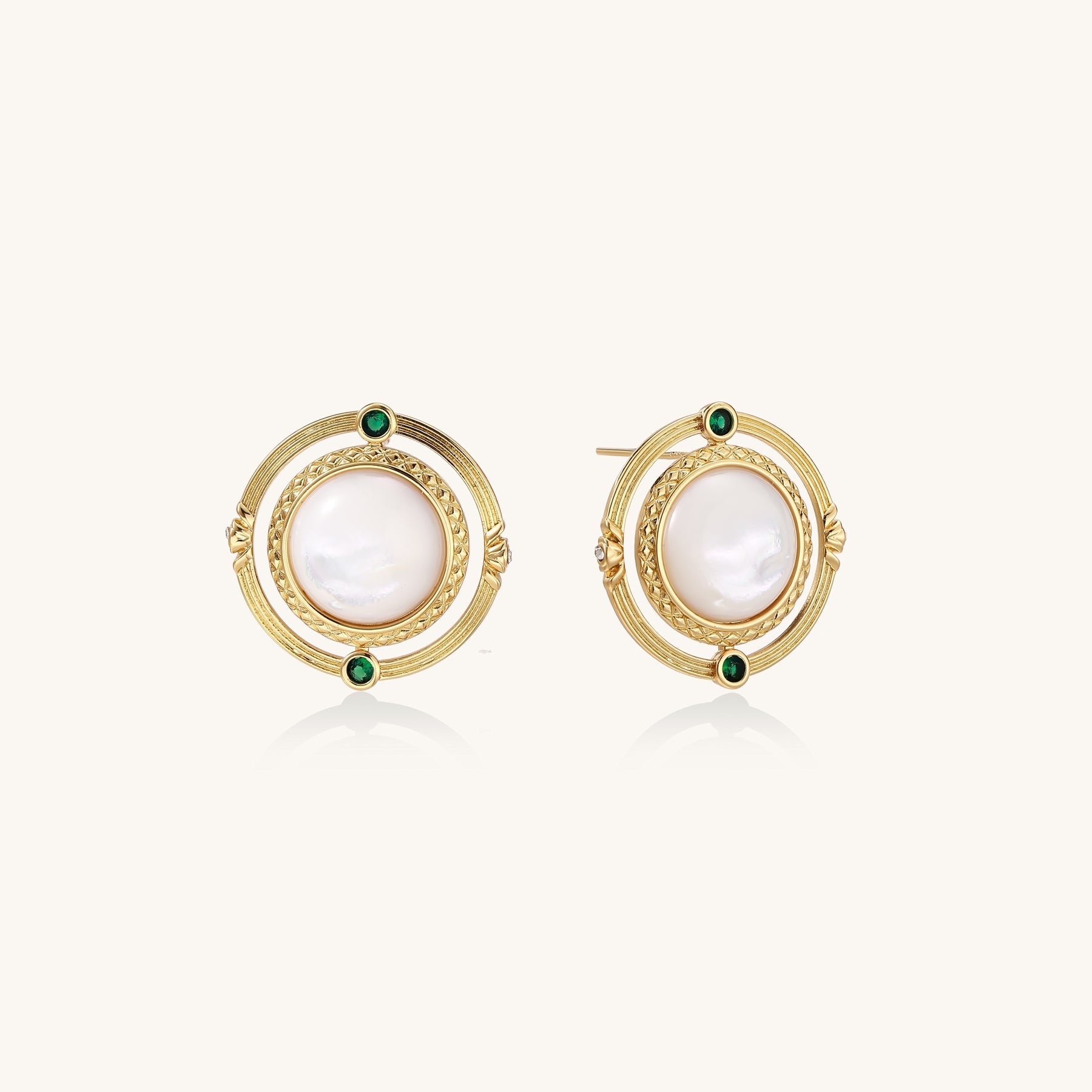 Close-up of gold-plated circular earrings with white mother-of-pearl centers and green zirconia accents on cream background.