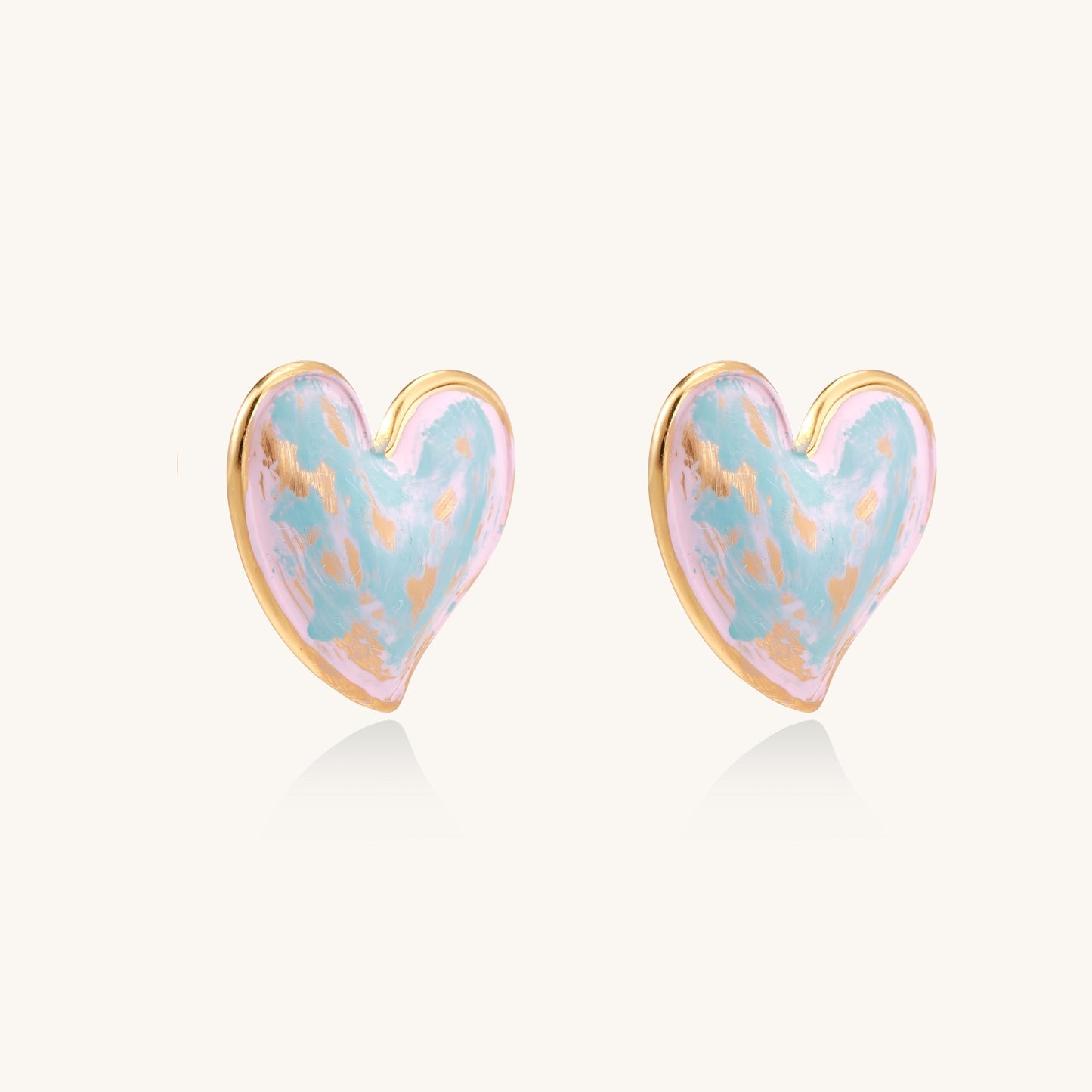 Heart-shaped stud earrings with iridescent blue-pink enamel finish and gold-plated brass frames against cream background