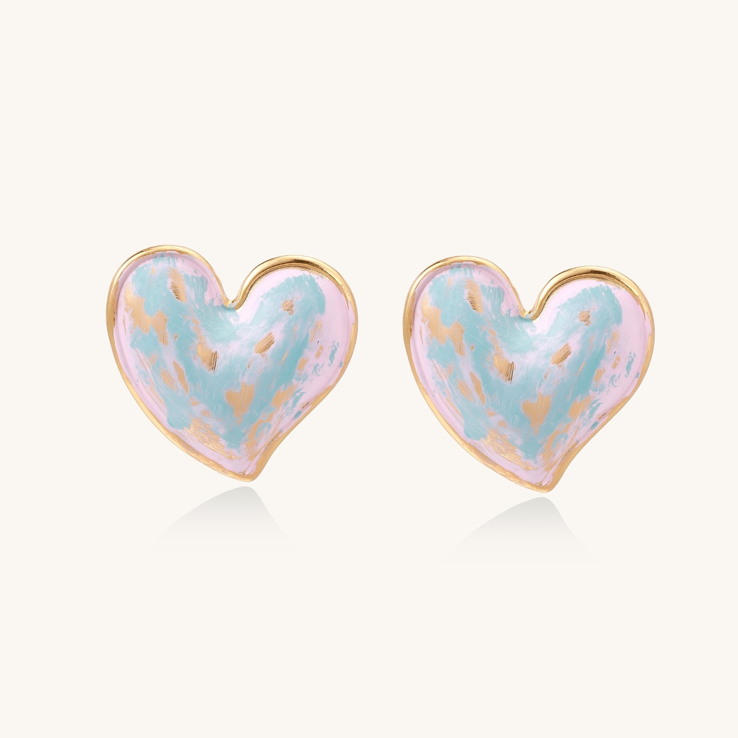 Heart-shaped stud earrings with iridescent blue-pink enamel finish and gold-plated brass frames against cream background