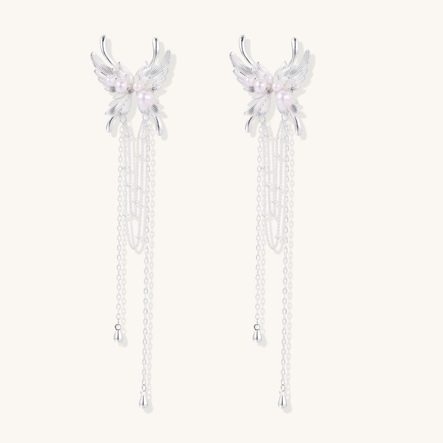 Elegant silver-plated drop earrings with wing-shaped tops, white pearl accents, and long chain tassels on white background.