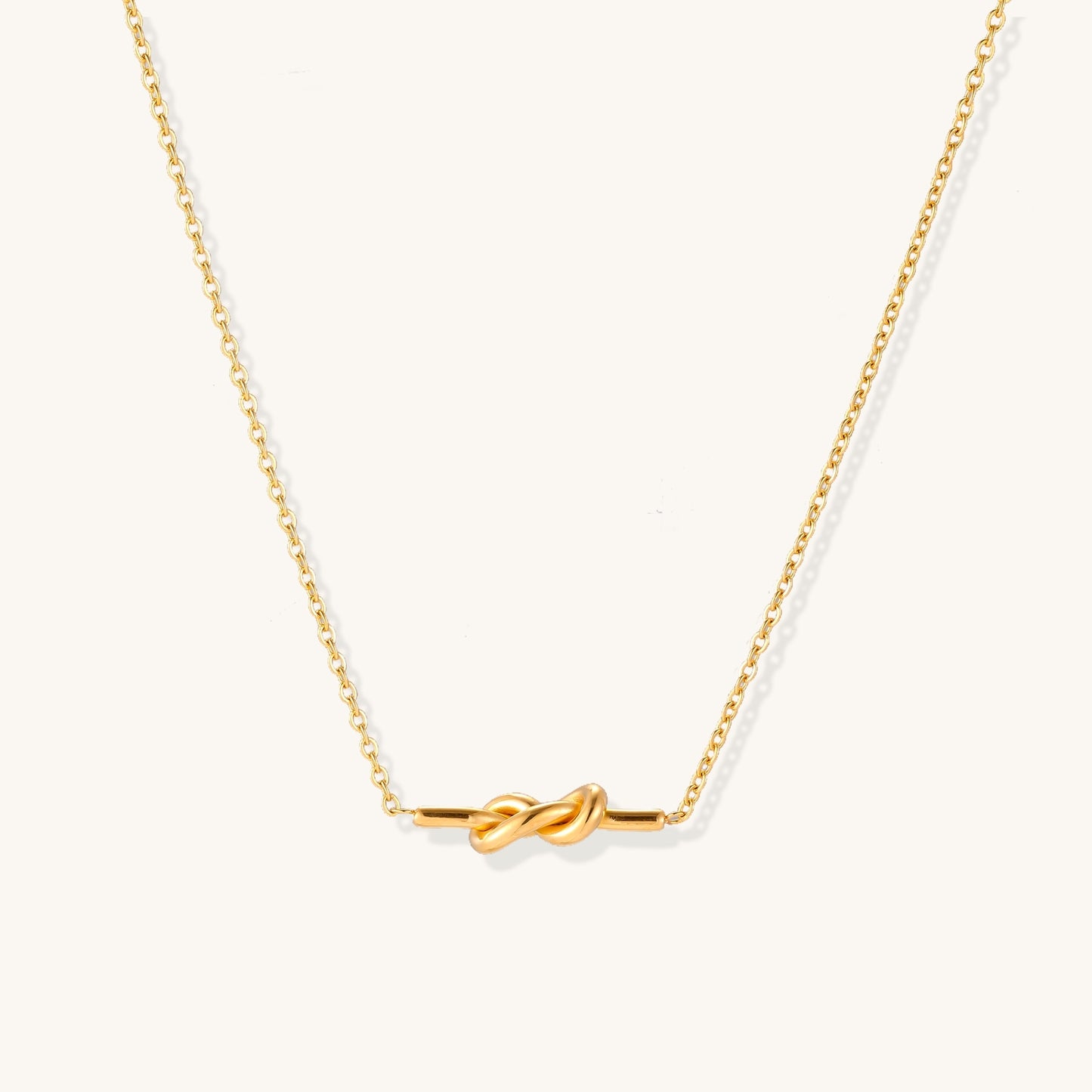 Dainty 18K gold plated stainless steel chain necklace with minimalist knot pendant on cream background.