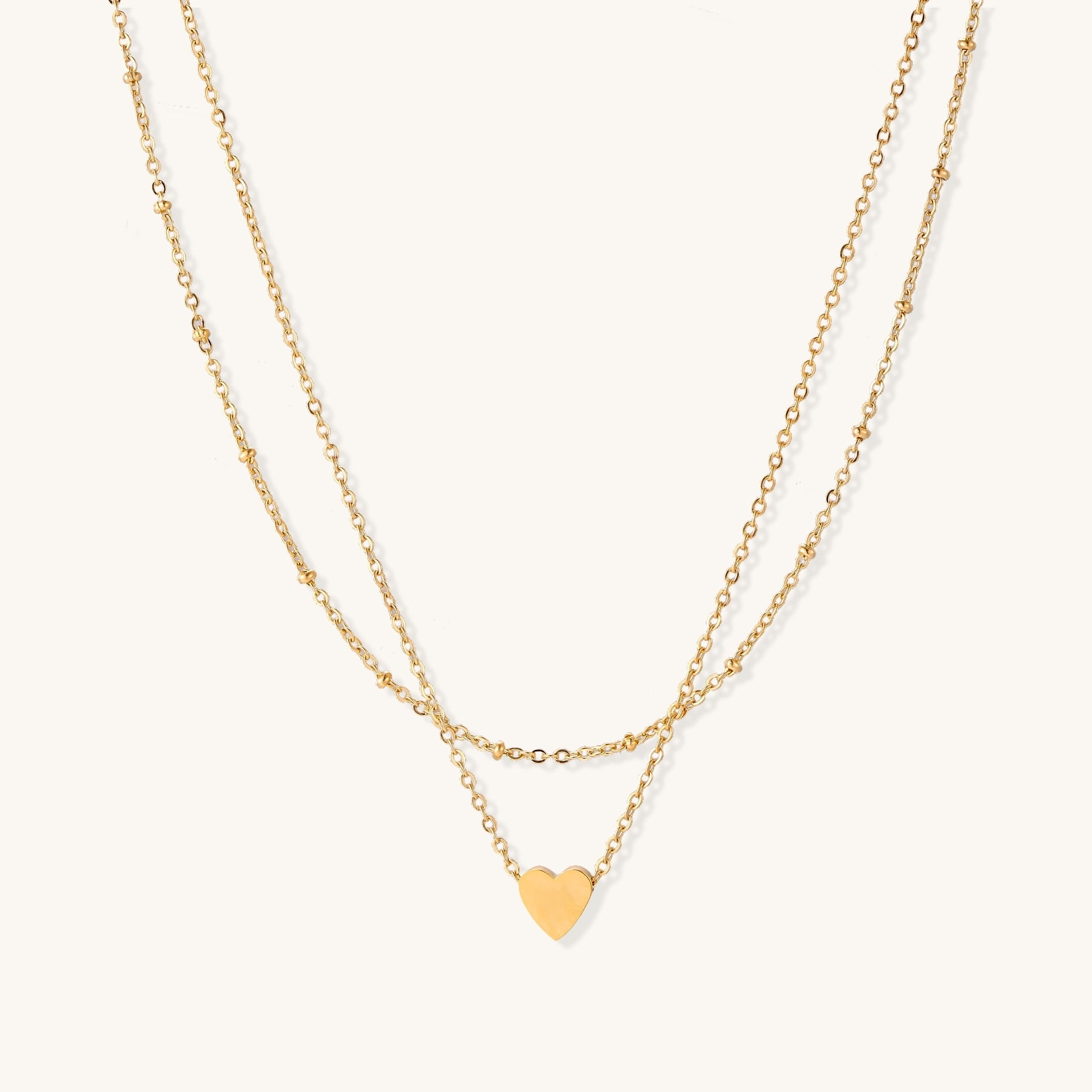 Two-layer gold-plated stainless steel necklace with small heart pendant displayed on beige background