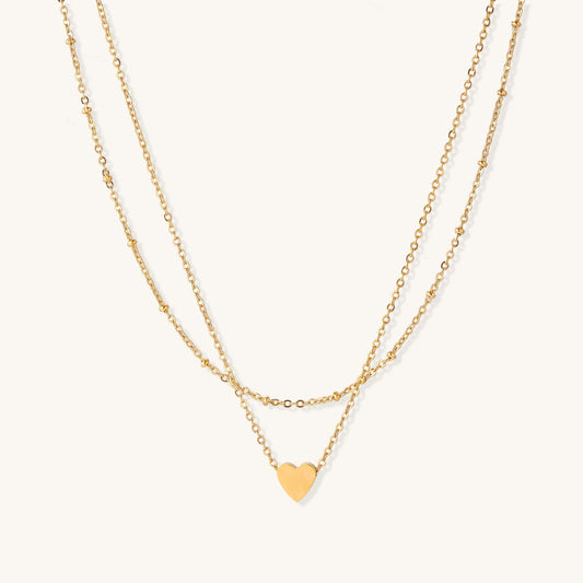 Two-layer gold-plated stainless steel necklace with small heart pendant displayed on beige background